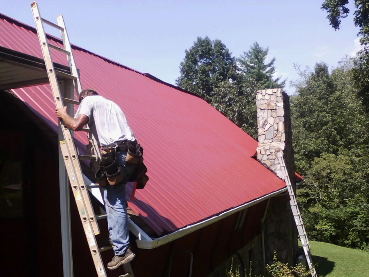 Roofer on a ladder beside a red metal roof and stone chimney — Soffit & Fascia Repair by Roofing Brothers in Wynot