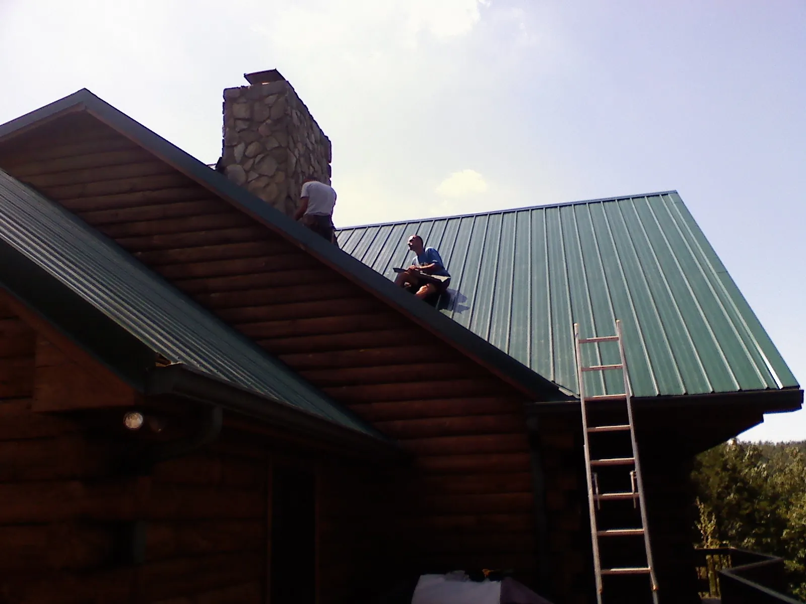 Roofers installing green metal panels beside a stone chimney on a log home in Wynot